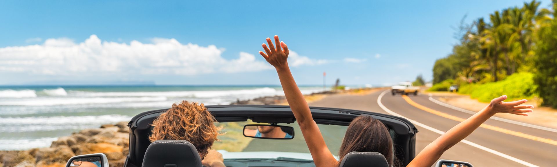 Zwei Personen im Auto mit offenem Verdeck, im Hintergrund kann man das Meer sehen