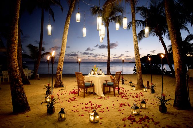 Ein gedeckter Tisch am Strand, Rosenblätter liegen am Boden und Laternen hängen im Baum
