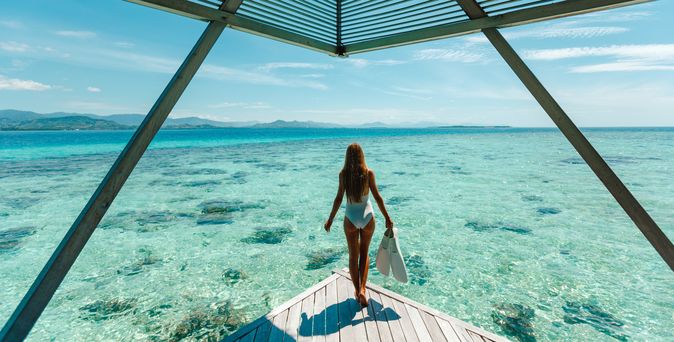 Eine Frau im Bikini steht am Ende eines Stegs und schaut aufs Meer