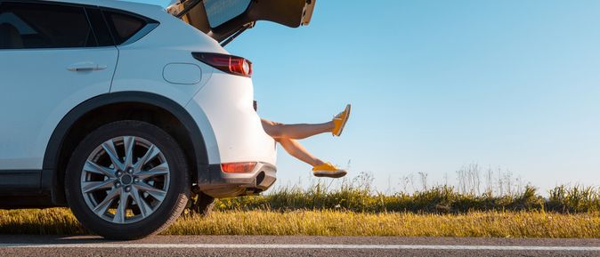 Ein Auto ist zur Hälfte von der Seite zu sehen und steht am Straßenrand an einer Wiese. Der Kofferraum ist auf und man sieht Beine herausragen