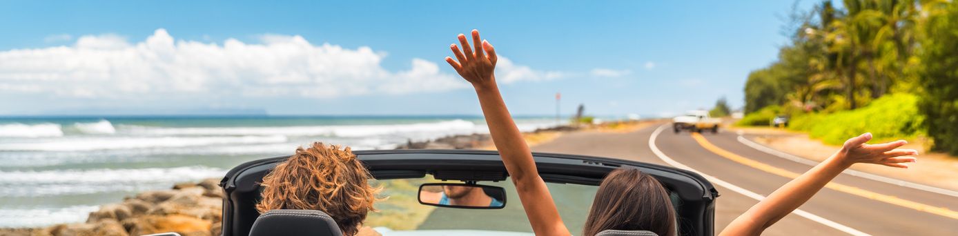 Zwei Personen im Auto mit offenem Verdeck, im Hintergrund kann man das Meer sehen