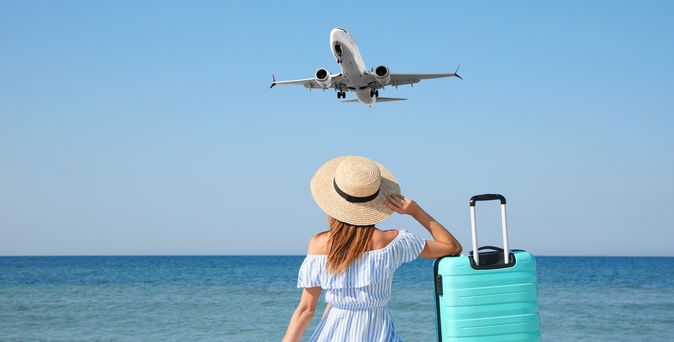 Frau im Sommerkleid und Sonnenhut sitzt am Meer und schaut auf das Meer. Ihr rechter Arm ist auf einen blauen Koffer gelehnt