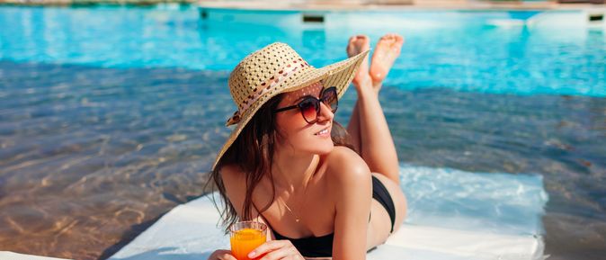 Eine lächelnde Frau im Bikini liegt auf einer Liege in einem Pool und schaut glücklich nach Rechts, in der Hand hält sie einen Orangen Cocktail