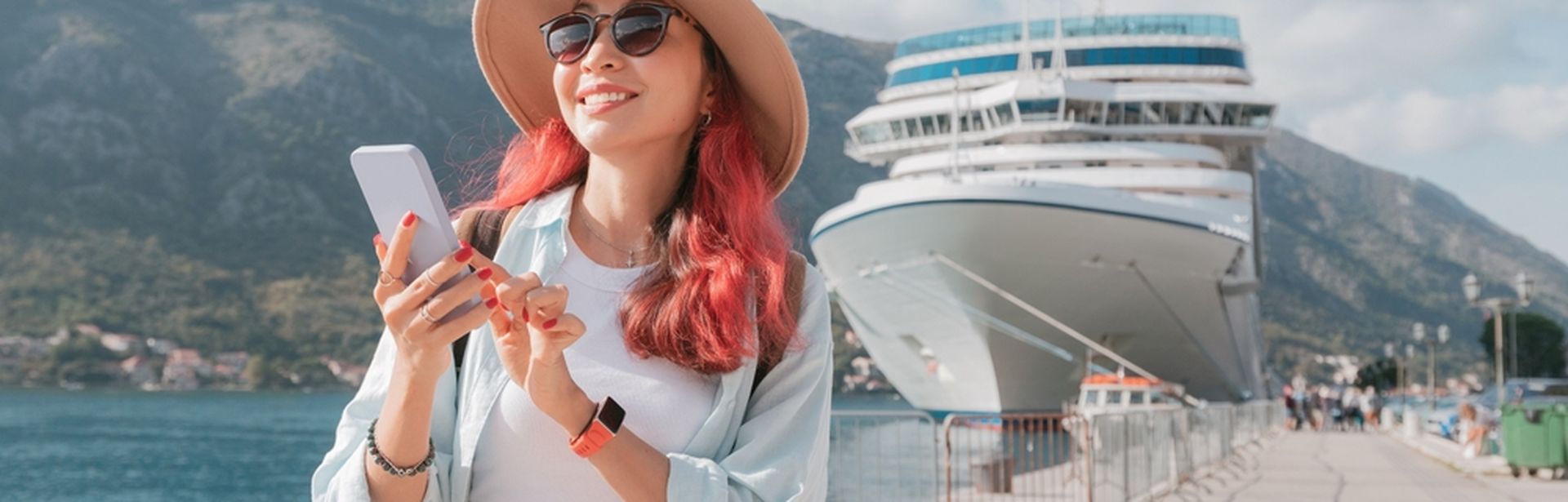 Eine Frau mit roten Haaren steht an einem Hafen, hält ihr Smartphone fest und lächelt; Im Hintergrund steht ein Kreuzfahrtschiff