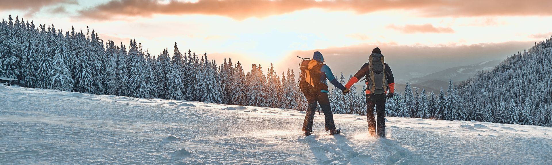 Zwei Personen stehen auf einem Schneebedeckten Berg und schauen Händchen haltend in den Sonnenuntergang