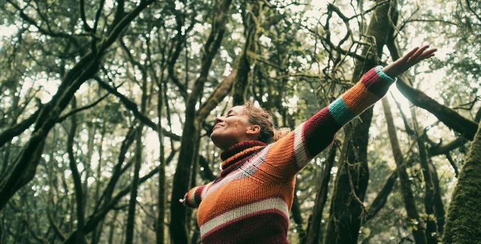 Eine Frau breitet glücklich die Arme aus und hat die Augen geschlossen, Sie steht in einem Wald voller dichter Bäume