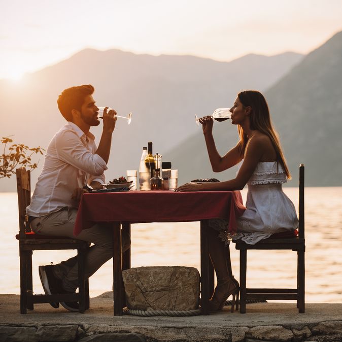 Eine Frau und ein Mann sitzen sich bei Sonnenuntergang an einem Tisch am Wasser gegenüber und trinken Wein