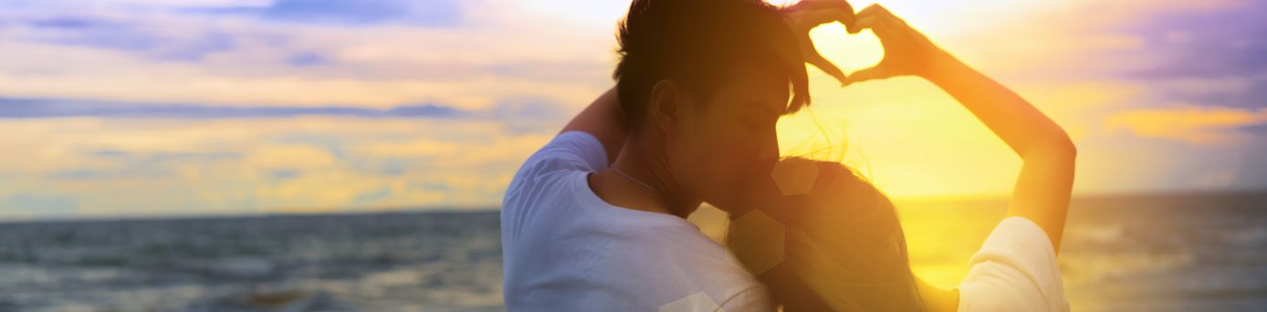 Ein Paar formt mit den Händen gemeinsam ein Herz und schaut bei Sonnenuntergang auf das Meer