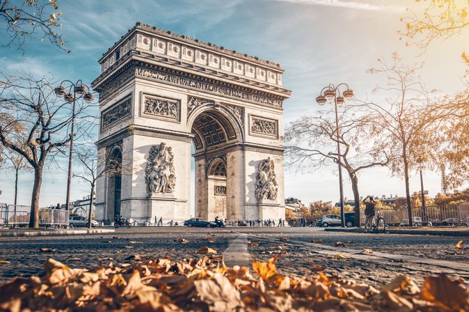 Der Arc de Triomphe im Herbst. im Vordergrund liegen braune Blätter auf dem Boden.