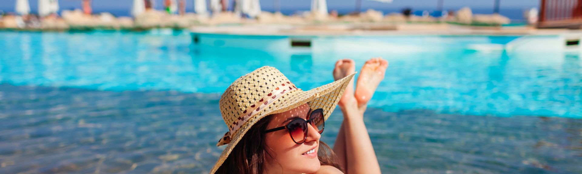 Eine lächelnde Frau im Bikini liegt auf einer Liege in einem Pool und schaut glücklich nach Rechts, in der Hand hält sie einen Orangen Cocktail