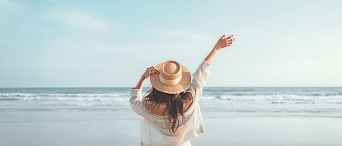 Eine Frau mit Hut von hinten, Sie schaut vom Strand aus auf das Meer und hebt den Arm 
