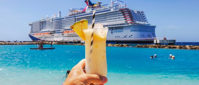 Eine Hand hält einen Cocktail hoch, im Hintergrund ist ein großes Kreuzfahrtschiff