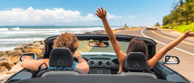 Zwei Personen im Auto mit offenem Verdeck, im Hintergrund kann man das Meer sehen