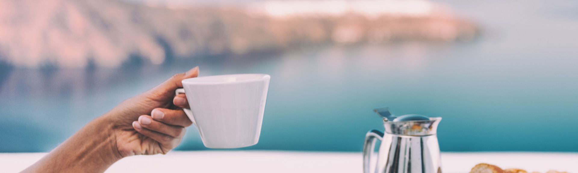 Ein gedeckter Tisch mit einer Kanne und Tellern mit Obst, eine Hand hält eine Tasse hoch, im Hintergrund ist das Meer