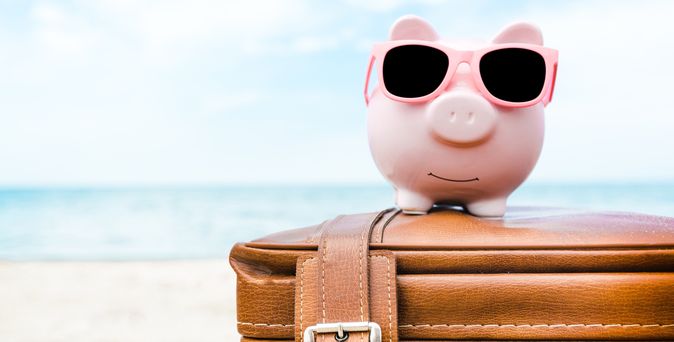 Ein rosanes Sparschwein mit Sonnenbrille sitzt auf einem Koffer am Strand