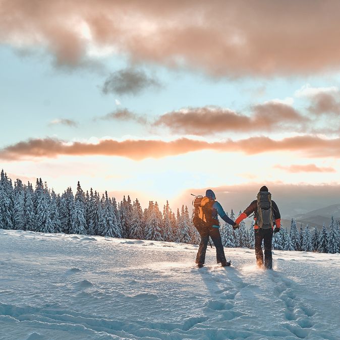 Zwei Personen stehen auf einem Schneebedeckten Berg und schauen Händchen haltend in den Sonnenuntergang