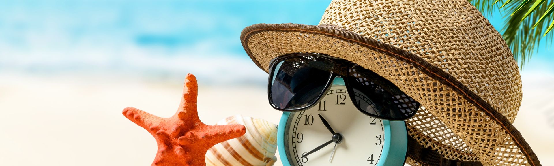 Ein Wecker mit Sonnenhut und einer Sonnenbrille liegen am Strand