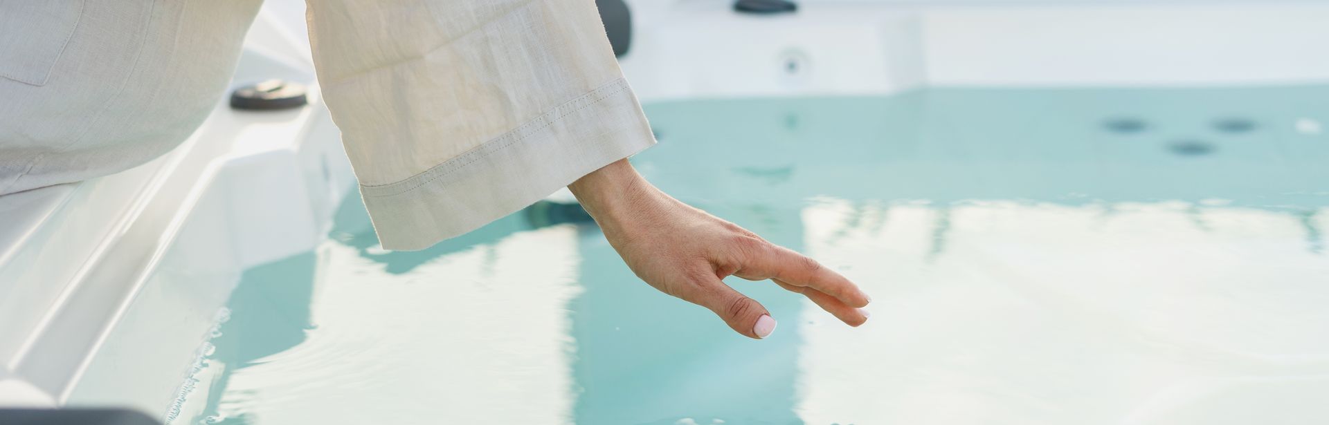 Eine Frau sitzt im Bademantel am Rand eines Whirlpools und hält Ihre Hand über die Wasseroberfläche