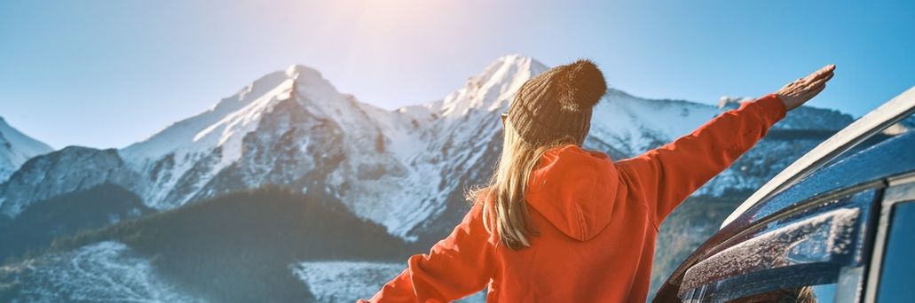 Frau steht an der offenen Tür eines Autos, blickt auf eine schneebedeckte Berglandschaft und streckt die Arme aus