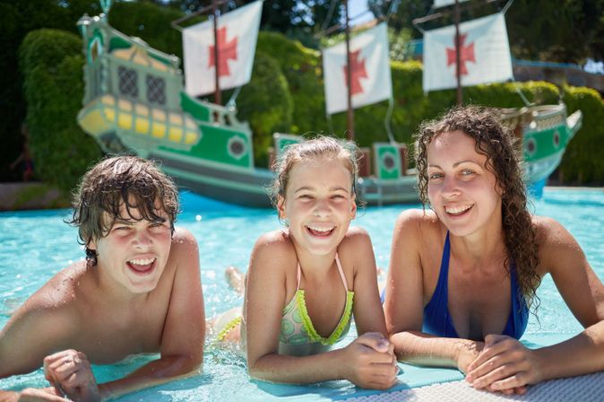 Zwei Mädchen und ein Junge lehnen am Rand eines Pools und lächeln in die Kamera, im Hintergrund sieht man ein Piratenschiff