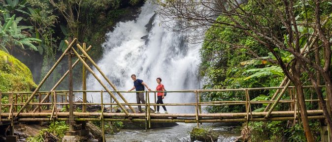 Eine Frau und ein Mann laufen Hand in Hand über eine Holzbrücke über einem Fluss, im Hintergrund ist ein großer Wasserfall