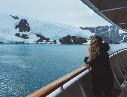 Eine Frau steht mit wehenden Haaren an der Reling eines Kreuzfahrtschiffes und schaut auf schneebedeckte Berge
