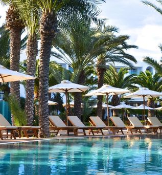 16:18Eleganter Hotelpool mit türkisblauem Wasser, gesäumt von hellen Holzliegestühlen und cremefarbenen Sonnenschirmen unter dicht gewachsenen Dattelpalmen, im Hintergrund die weiße Hotelfassade.