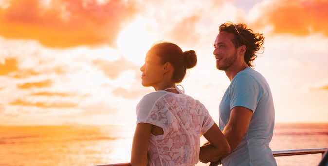 Eine Frau und ein Mann stehen bei Sonnenuntergang an der Reling eines Kreuzfahrtschiffes