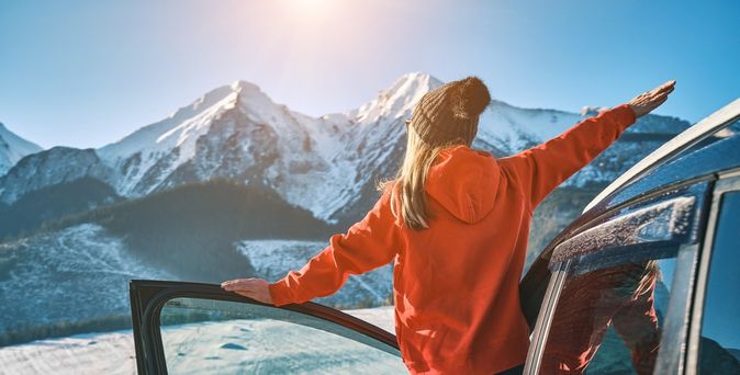 Frau steht an der offenen Tür eines Autos, blickt auf eine schneebedeckte Berglandschaft und streckt die Arme aus