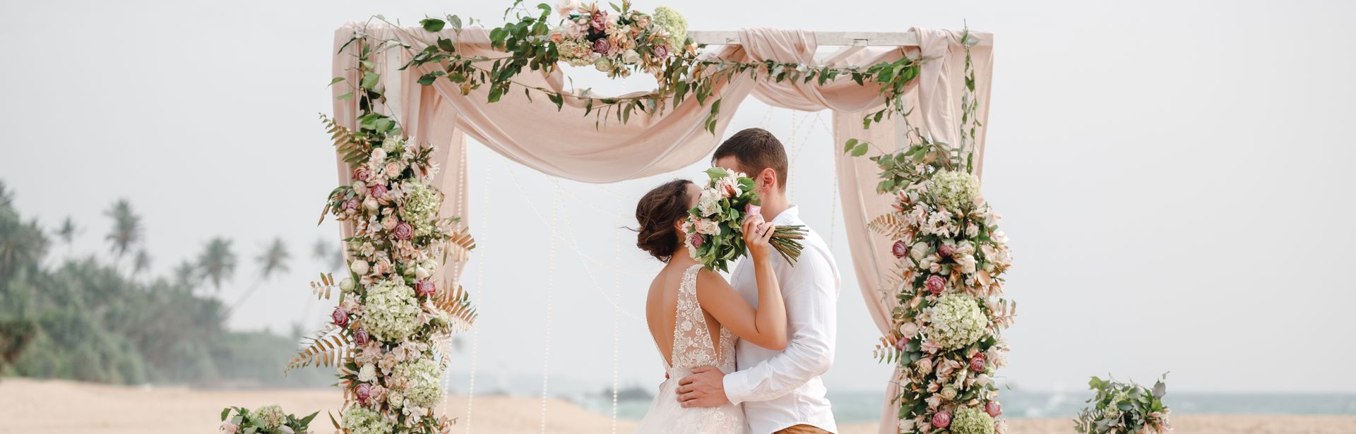Ein Hochzeitspaar küsst sich unter einem Traubogen am Strand