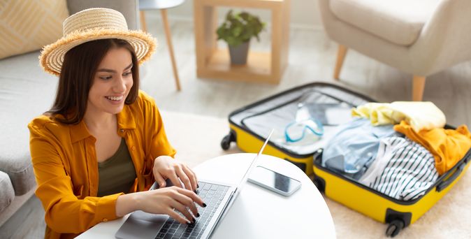 Eine sich freuende Frau tippt an einem Laptop. Im Hintergrund liegt ein gepackter Koffer