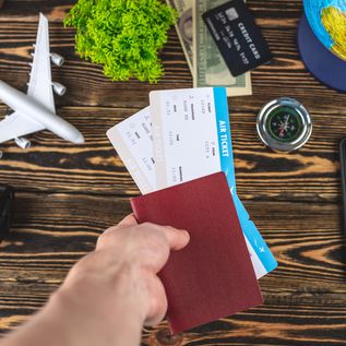 Auf einem Holztisch liegen ein Handy, ein Miniaturflugzeug, eine Kamera, ein Kompass, Geld und ein Globus. Eine Hand hält einen Reisepass und zwei Flugtickets