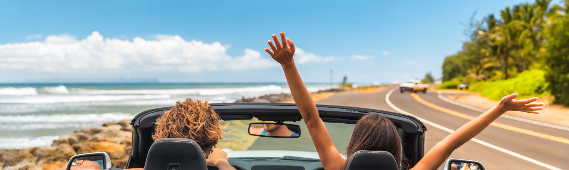 Zwei Personen im Auto mit offenem Verdeck, im Hintergrund kann man das Meer sehen
