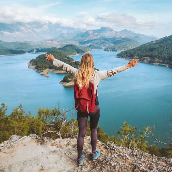Eine Frau mit Rucksack schaut von einer Klippe auf einen großen See, eine Berglandschaft und breitet die Arme aus
