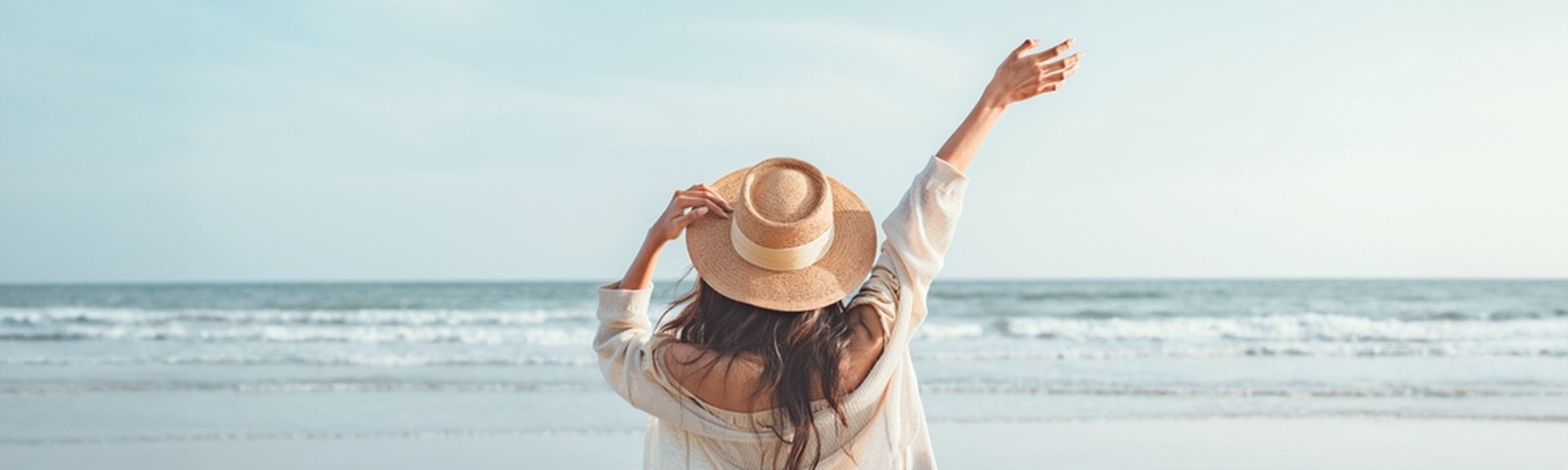 Eine Frau mit Hut von hinten, Sie schaut vom Strand aus auf das Meer und hebt den Arm 