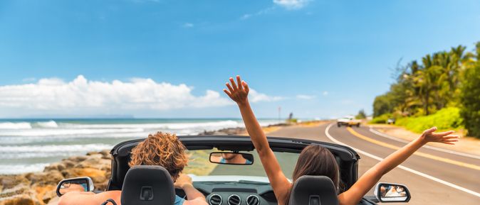 Zwei Personen im Auto mit offenem Verdeck, im Hintergrund kann man das Meer sehen