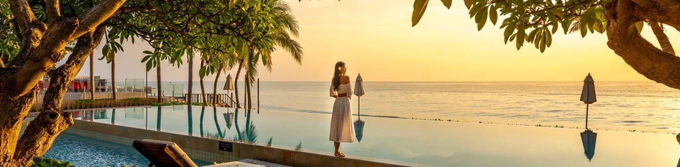 Frau steht bei Sonnenuntergang vor einem Pool der direkt vor einem Meer liegt
