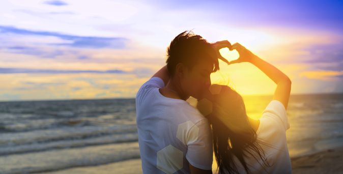 Ein Paar formt mit den Händen gemeinsam ein Herz und schaut bei Sonnenuntergang auf das Meer