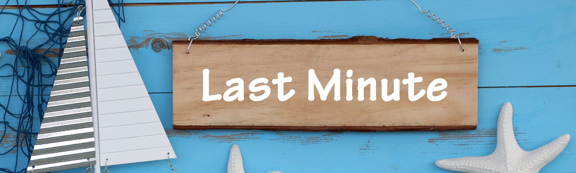 Eine blaue Holzwand mit einem Schild mit der Aufschrift "Last Minute", davor stehen zwei Seesterne, ein kleines Holzboot und eine kleine Flasche mit Muscheln und Sand 