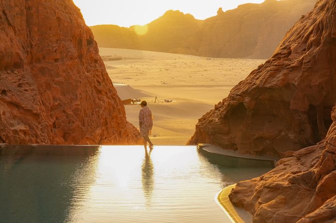 Person läuft am Rand eines Infinity Pools entlang, der zwischen Felsen steht. Im Hintergrund ist Sand und ein Sonnenuntergang zu sehen