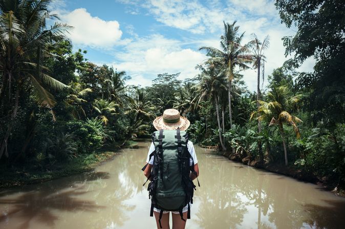 Frau mit Strohhut und Rucksack schaut auf Wasser und Palmen
