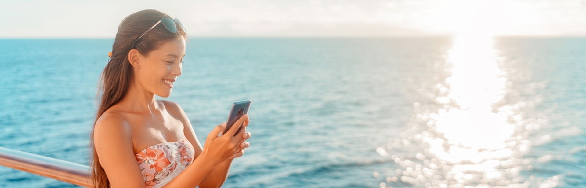 Eine Frau steht lächelnd an der Reling eines Kreuzfahrtschiffes und schaut auf ihr Smartphone