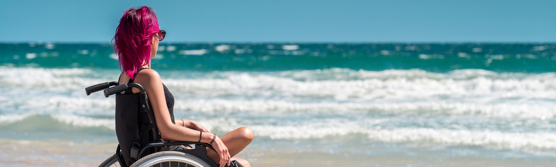 Frau im Rollstuhl mit einem Bein sitzt entspannt am Strand und blickt aufs Meer