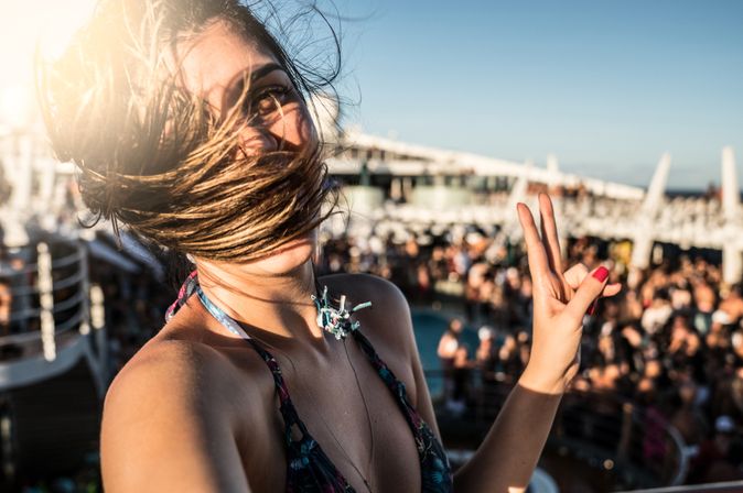 Eine Frau macht auf einem Kreuzfahrtschiff ein Selfie, Ihre Haare wehen im Wind und im Hintergrund sieht man eine feiernde Menge