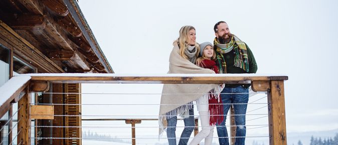 Eine Familie steht in Decken gehüllt auf dem Balkon Ihres Hotels im Schnee