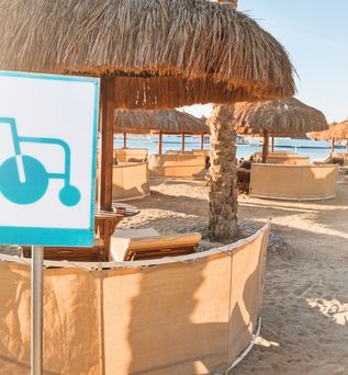 Strand mit mehreren Tischen und Schirmen, ein Schild im Vordergrund auf dem ein Rollstuhl abgebildet ist