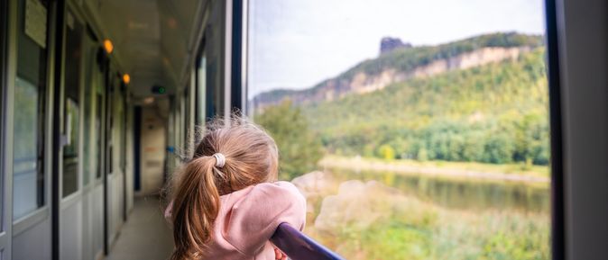 Ein Kind steht am Fenster in einem Zug und schaut auf die grüne Landschaft