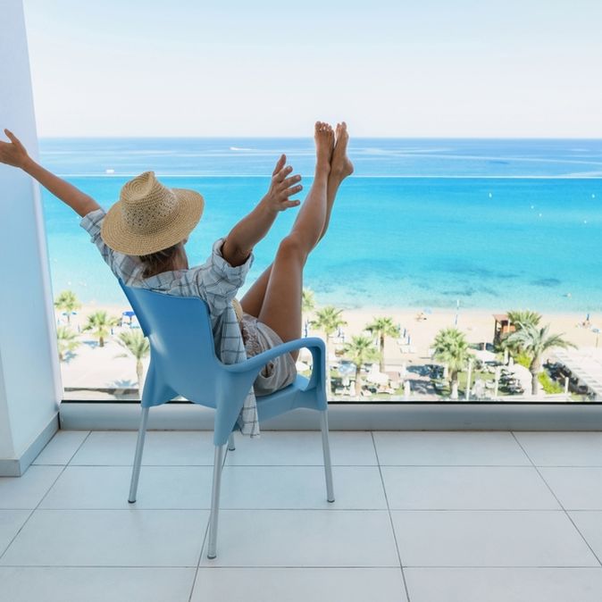 Eine Frau in einem Hemd und einem Strohhut sitzt auf dem Balkon Ihres Hotelzimmers und streckt Arme und Beine in die Luft