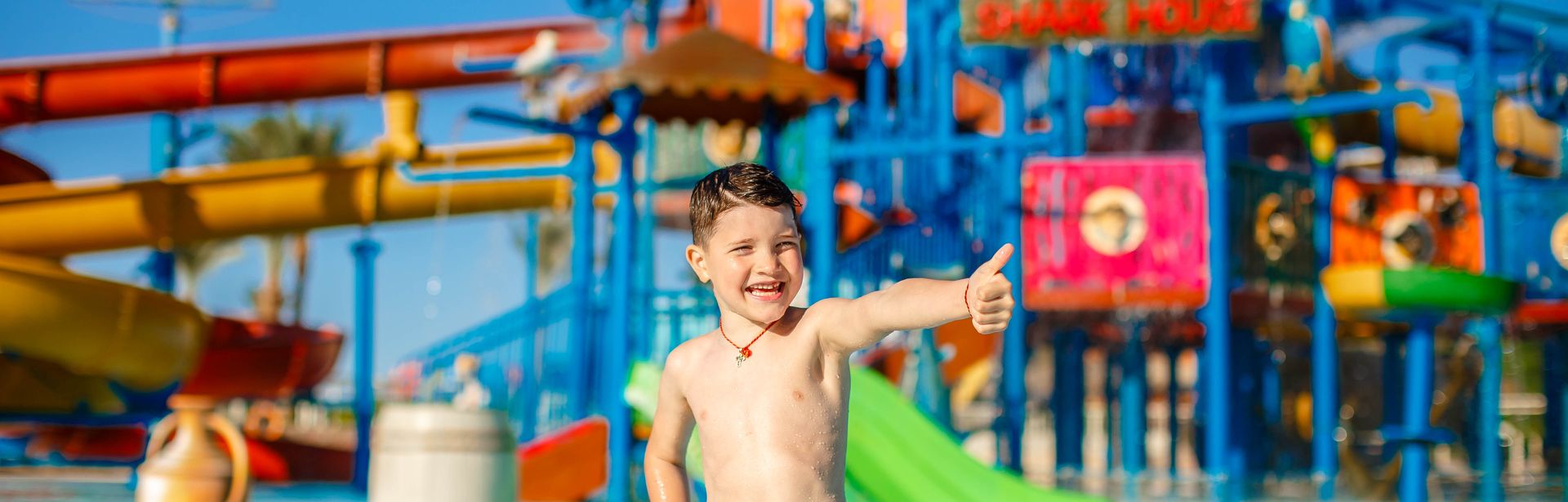 Ein Junge in einer gelben Badehosen steht vor einem großen Aquapark und zeigt einen Daumen nach oben
