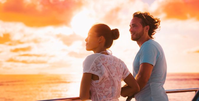 Eine Frau und ein Mann stehen bei Sonnenuntergang an der Reling eines Kreuzfahrtschiffes